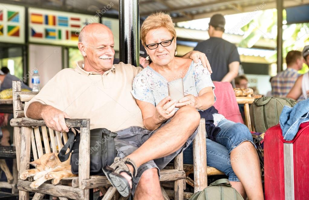 Happy senior couple sitting and using mobile smart phone with travel suitcase during adventure trip around the world - Active elderly lifestyle concept and interaction with new trends and technology