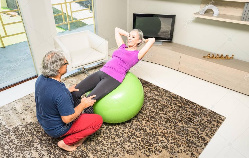 Active elderly couple training with swiss ball at home - Retired people at exercise fitness activity in gym living room - Mature man coaching lady woman on crunch sit ups abdominal sport execution