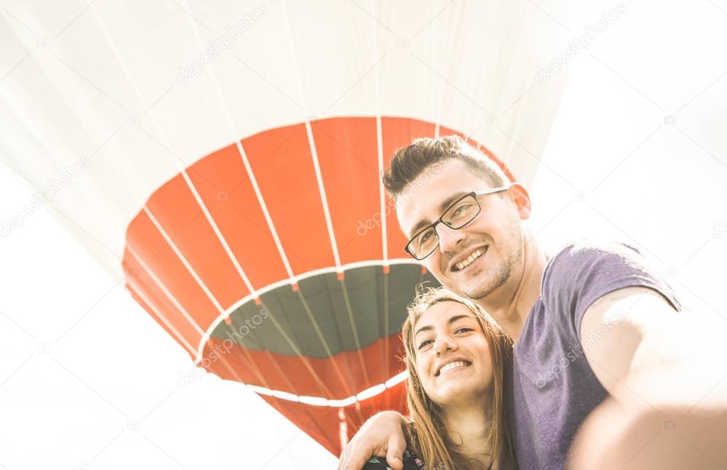 Happy couple in love on honeymoon vacation taking selfie at hot air balloon - Summer travel concept with young people travelers having fun at excursion - Vintage contrast retro filter with backlight