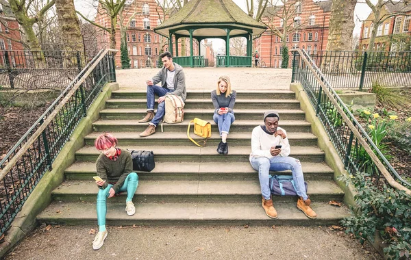 Группа многорасовых друзей, использующих мобильный смартфон, сидящий на лестнице в парке - Молодые хипстеры, зависимые от смартфона в сообществе социальных сетей - Технологическая концепция - Обесцвеченные тона — стоковое фото