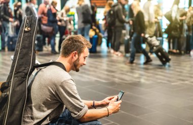 Adam ve gitar sırt çantası - yüz odaklanmak ile mobil akıllı telefon - terminal kapısında bekleyen uçak için avare kişi - Yolculuk tutkusu seyahat gezi kavramı kullanarak Uluslararası Havaalanı'nda maceracı adam