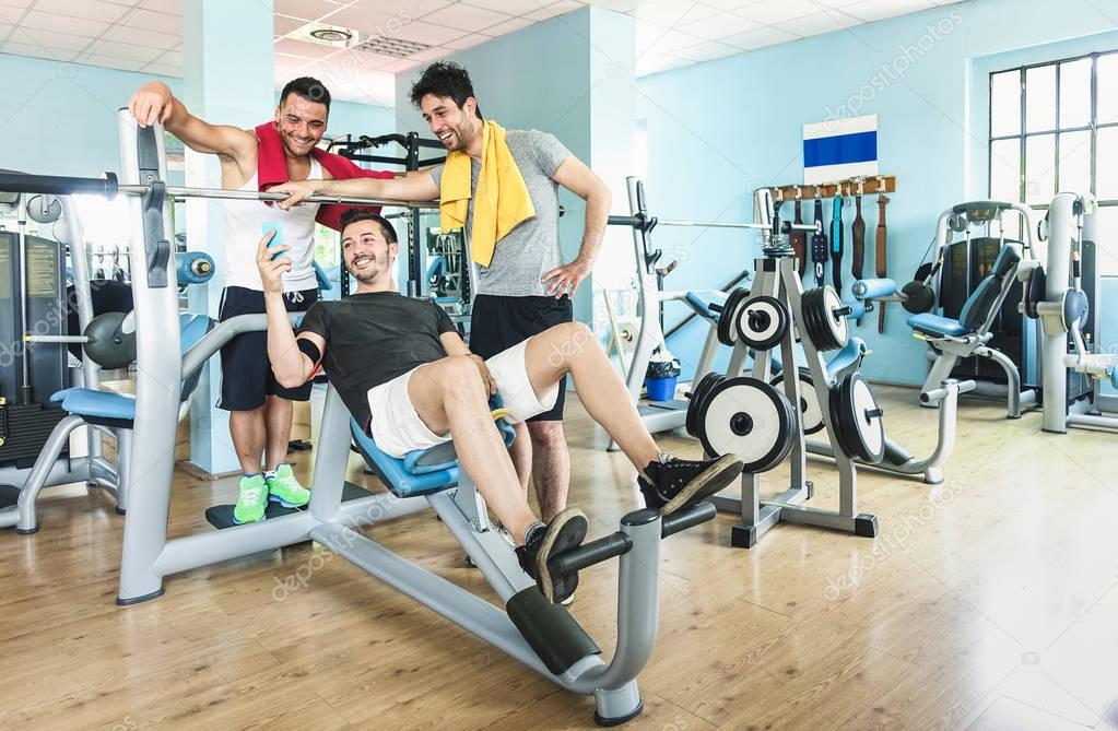 Group of sportive friends using mobile phone at gym fitness club - Happy sporty people in weight room training - Social gathering concept in sport lifestyle context - Main focus on guys interaction