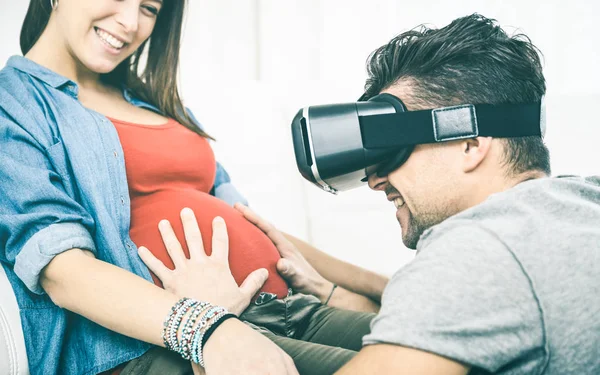 Young couple with pregnant woman having fun with vr goggles tech headset - Happy people with man husband using virtual reality device on wife belly - Wearable technology concept - Clear vintage filter