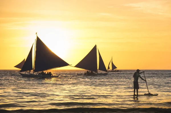 Silhouette of sailing boats at sunset in Boracay island - Unrecognizable man with paddle surfing - Exclusive travel destination in Philippines - Warm vintage filtered look - Focus in foreground — Stock Photo, Image