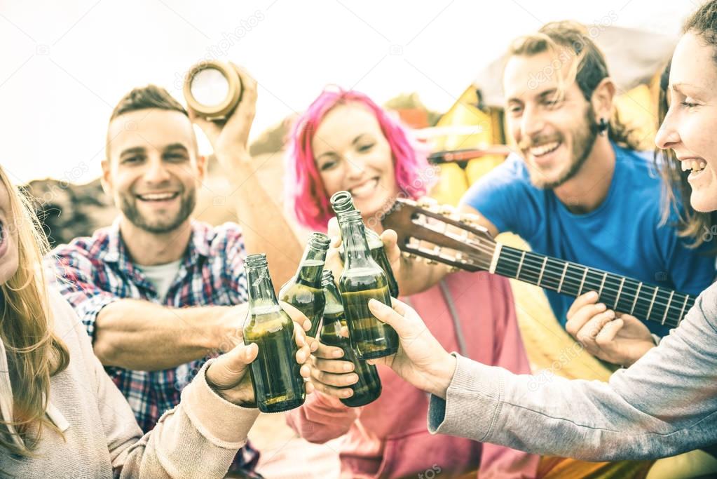 Young friends having fun together at beach camping party - Friendship travel concept with hipster people wanderers playing guitar and drinking bottled beer at summer surf camp - Warm sunshine filter