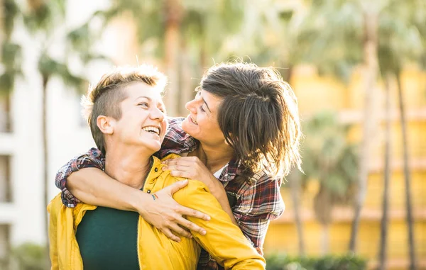 Felices novias juguetonas enamoradas compartiendo tiempo juntas en viaje de viaje en abrazo a cuestas - Concepto de amistad de las mujeres con las niñas pareja divirtiéndose en ropa de moda al aire libre - Filtro de puesta de sol cálido brillante —  Fotos de Stock