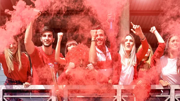 Jóvenes aficionados al fútbol animando con humo de colores viendo el partido de fútbol juntos en el estadio - Amigos grupo de personas con camisetas rojas que se divierten en el concepto de campeonato mundial de deporte — Foto de Stock