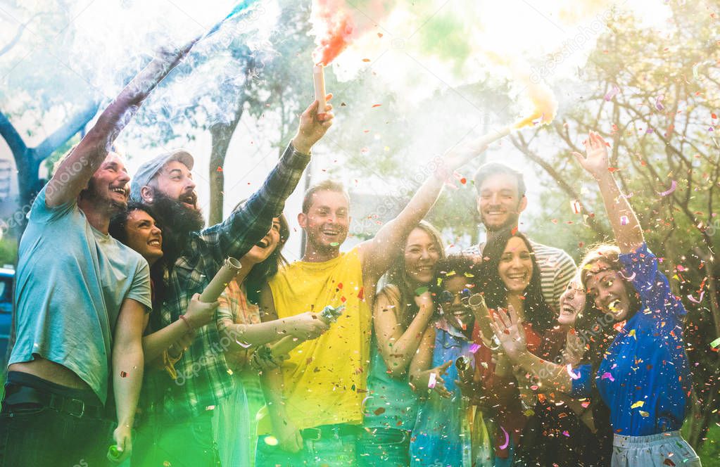 Happy friends having fun at garden party with multicolored smoke bombs outdoor - Young millennial students celebrating spring break together - Genuine youth concept - Focus on confetti and left people