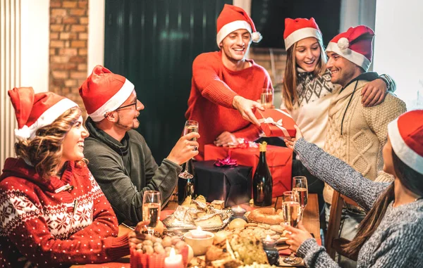 กลุ่มเพื่อนสวมหมวกซานตาให้ของขวัญคริสต์มาสกันและกัน ขนมปังแชมเปญไวน์ที่บ้านเอ็กซ์มาสมื้อค่ํา แนวคิดวันหยุดกับคนหนุ่มสาวแบ่งปันเวลาด้วยกันและสนุกสนานในฤดูหนาว — ภาพถ่ายสต็อก