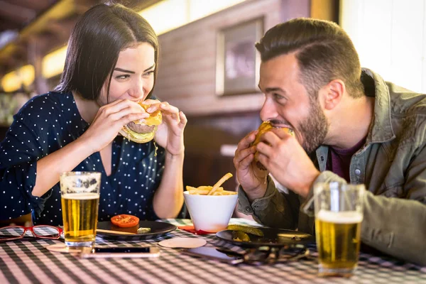 Pasangan bahagia bersenang-senang makan burger di pub restoran cepat saji orang-orang muda menikmati momen di lokasi restoran dalam ruangan konsep hubungan dengan wajah perempuan dengan filter hidup yang hangat — Stok Foto