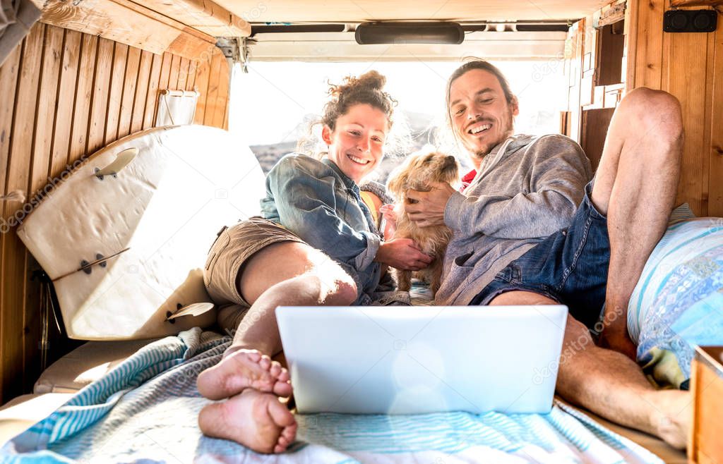 Digital nomad couple with cute dog using laptop on retro mini van transport - Travel life inspiration concept with indie people on minivan adventure trip watching pc on relax moment - Warm filter