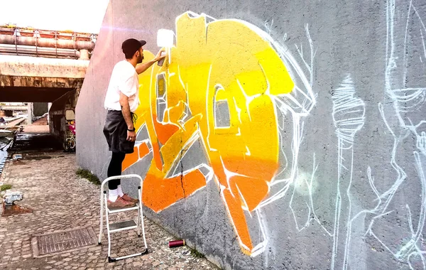 パブリックスペースの壁で色の落書きに取り組むストリートアーティスト-現代美術は黄色とオレンジのエアロゾル色スプレーでライブ壁画を描く都市の男の概念を実行します-明るいサンフレアフィルター — ストック写真