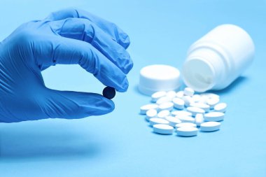 Beyaz haplar ve elinde mavi eldivenli siyah bir hap. Farklı haplar. Fotokopi alanına sahip açık mavi kağıt arkaplanı tıbbi ve farklı kavramları.