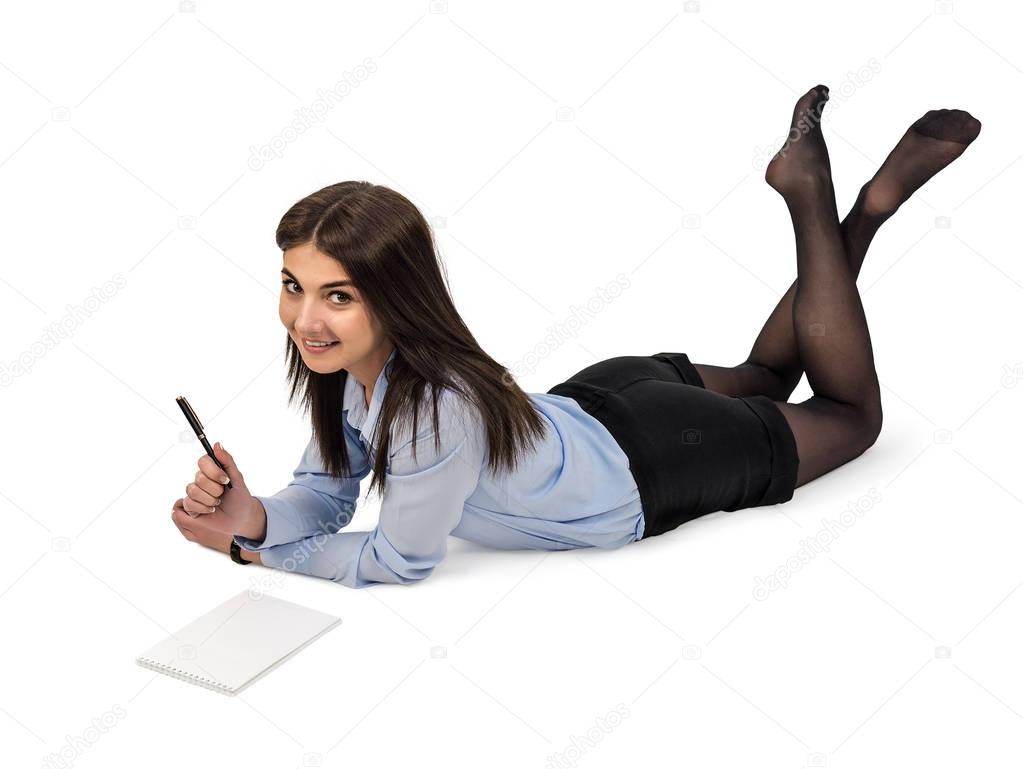 Cheerful young woman with a ballpoint pen and a notepad is lying on the floor. White isolated background.    
