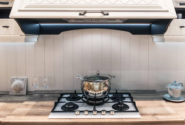 A clean, shiny metal saucepan stands on a gas stove. The kitchen is in classical style with a wooden tabletop and a large modern cooker hood.