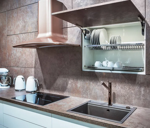 Clean, modern kitchen in Loft style. An induction hob and a metal sink are built into the marble countertop. A large hood and an open shelf for drying dishes are built into the kitchen cabinet, lined with stone.