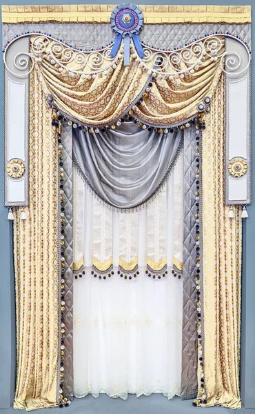 Luxurious interior decoration in the palace style. Curtains made of fabric of gold color with neutral gray inserts. A multi-tier figured pelmet, decorated with rhinestones, fringed and satin ribbons. — Stock Photo, Image