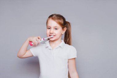 Küçük tatlı kız gülümser ve dişlerini gri bir arka planda renkli bir fırçayla fırçalar. Diş hijyeni. Mutlu küçük kız dişlerini fırçalıyor.