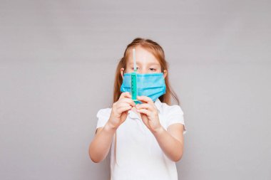 Gri arka planda, elinde şırınga ve tıbbi maske olan küçük bir kız.