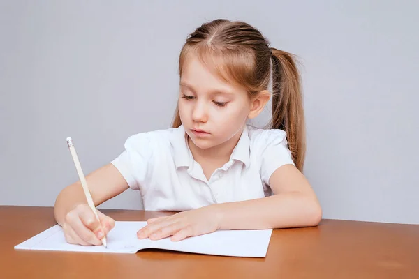 Uma menina de 6 anos senta-se a uma mesa com uma camiseta branca de manga curta e faz trabalhos de casa com um lápis nas mãos. . — Fotografia de Stock