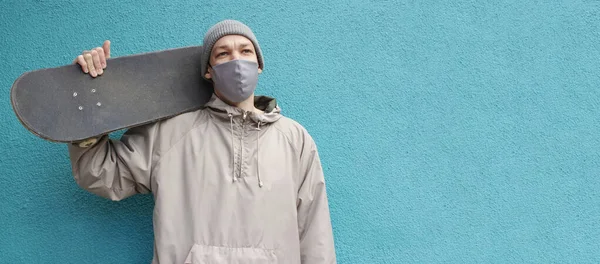 Yüz maskeli Avrupalı adam kaykayı omuzunda tutuyor. Turkuaz arka plan. Web sitesi ya da sosyal ağlar için yatay pankart. Mesaj için yer var. Şapkalı bir kaykaycı. Coronavirus koruma tedbirleri.