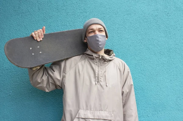 Tıbbi maskeli Avrupalı adam kaykayı omzunda tutuyor. Doymuş turkuaz arka plan. Gri ceketli ve şapkalı bir kaykaycı. Coronavirus koruma tedbirleri.