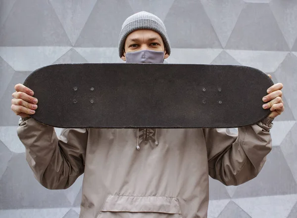 Europeisk kille i medicinsk mask håller skateboard på axeln. Grå betong vägg bakgrund. Skateboardåkare i grå jacka och hatt. Skyddsåtgärder mot coronavirus. — Stockfoto