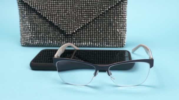 Damska torba na sprzęgło, telefon i okulary na niebieskim tle. — Wideo stockowe