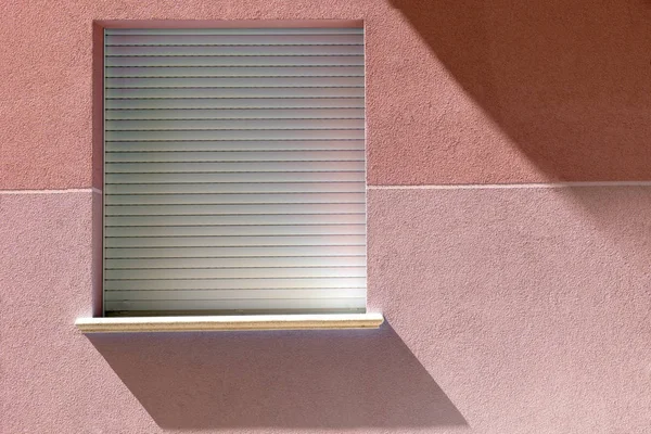 Minimalist blue window with shadow from the sun on a wall. A square red window hanging on a side wall of house