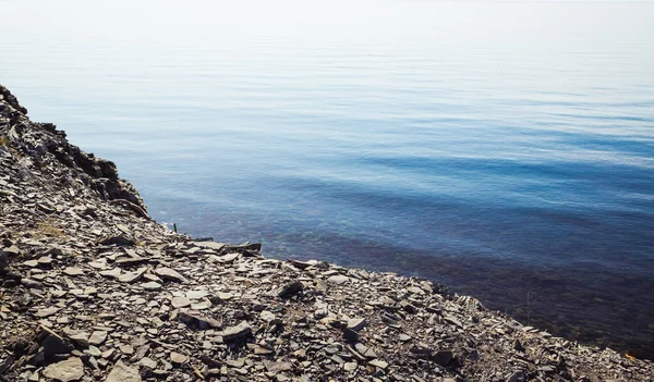 Blue calm sea under a rocky mountain cliff with sharp flat stones