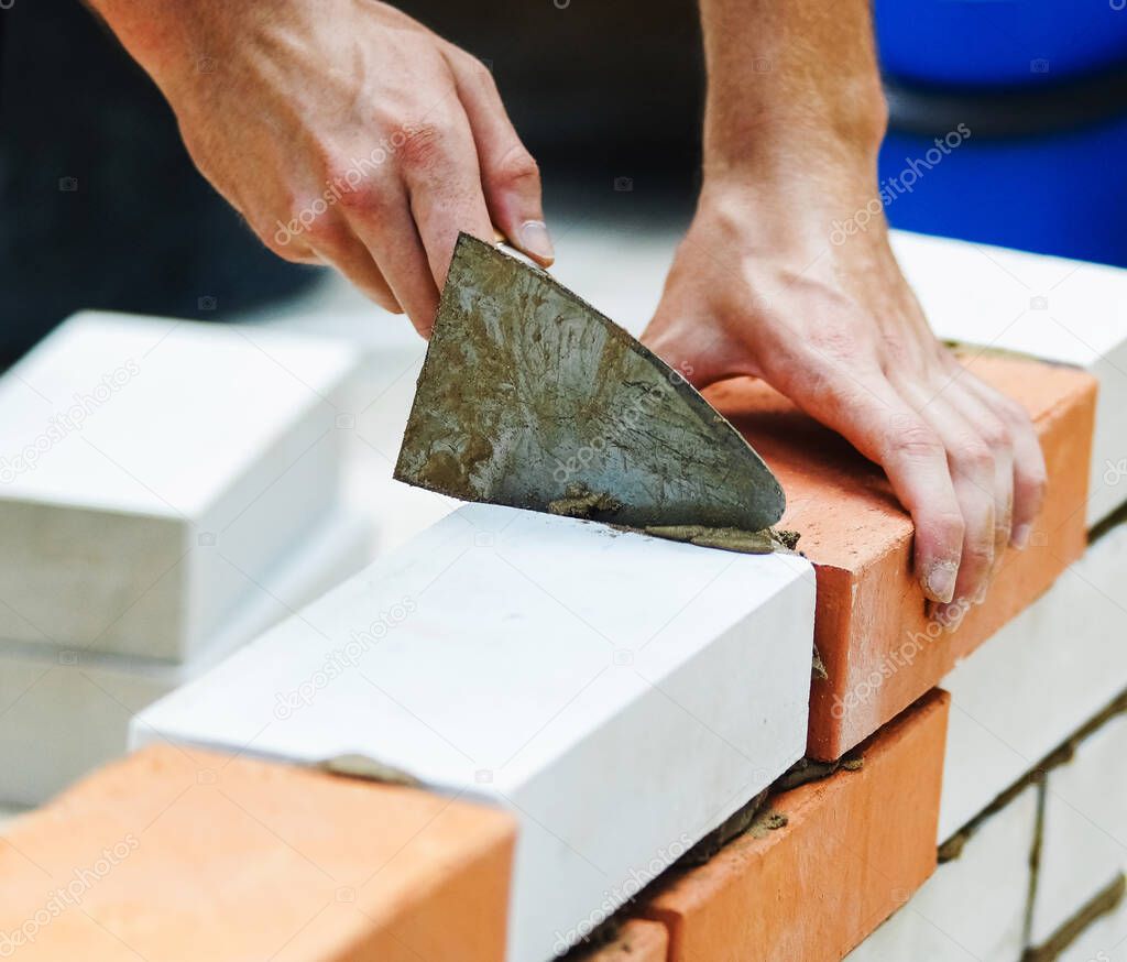 The builder puts cement with a trowel in the gap between the bricks. Brick wall made of white and orange bricks.