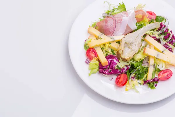 Dans une assiette blanche, mélanger la salade avec les légumes frais et le fromage. salade de tomates cerises, laitue, artichaut mariné, oignon, fromage, jambon — Photo