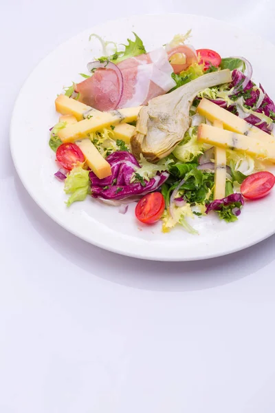 Mezclen la ensalada con hortalizas frescas y el queso en el plato blanco. ensalada de tomate cherry, lechuga, alcachofa marinada, cebolla, queso, jamón —  Fotos de Stock