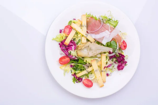 Salat mit frischem Gemüse und Käse auf einem weißen Teller. Kirschtomatensalat, Salat, marinierte Artischocke, Zwiebeln, Käse, Schinken — Stockfoto