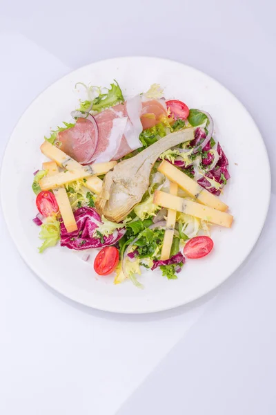 Dans une assiette blanche, mélanger la salade avec les légumes frais et le fromage. salade de tomates cerises, laitue, artichaut mariné, oignon, fromage, jambon — Photo