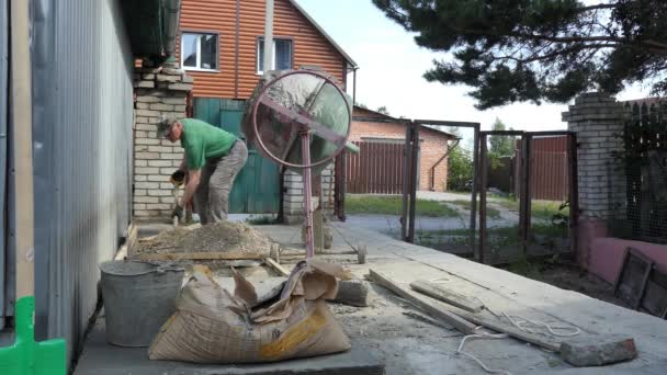 混凝土在家里起作用 一个人把一铲沙子和砾石扔进旋转的混凝土搅拌机 — 图库视频影像