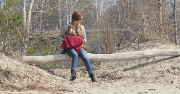 一个穿着牛仔裤 自我隔离的女人坐在沙滩上的圆木上 旁边是尚未被树叶覆盖的春树 她环顾四周 — 图库视频影像