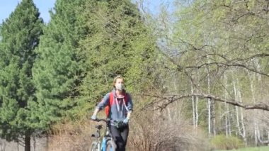 Kendini soyutlama. Tıbbi maske takan bir kadın bisikletini Sibirya sedir ağaçlarının arasındaki parkta yaya olarak sürüyor. Yakınlaştırılıyor.