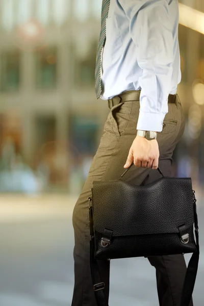 Uomo d'affari che cammina e tiene in mano una valigetta di pelle. Città moderna alle spalle — Foto Stock
