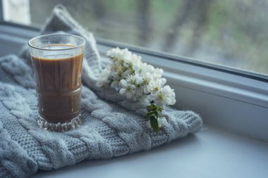 Kompozisyon, sütle birlikte bir fincan aromatik kahve, bir tüvit çiçek ve pencereye örülmüş yün bir eşarptan oluşuyor. Gündüz fotoğrafında.