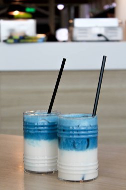 İki bardak buzlu bezelye latte. Mavi çay katmanlı, sütlü, ahşap masada siyah saman ve buzlu kokteyl.