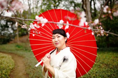 Beyaz kimono giyen Japon bir kadın, Tokyo, Japonya 'da kiraz çiçeklerinin açmasını izlerken kırmızı bir şemsiye tutuyor..