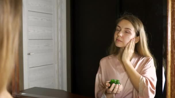 Bella donna sorridente che applica la crema alla pelle e si guarda allo specchio in camera da letto — Video Stock