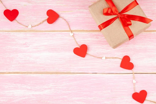 Pudełko z czerwoną wstążką i papierowe serca na sznurze juty na różowym tle — Zdjęcie stockowe
