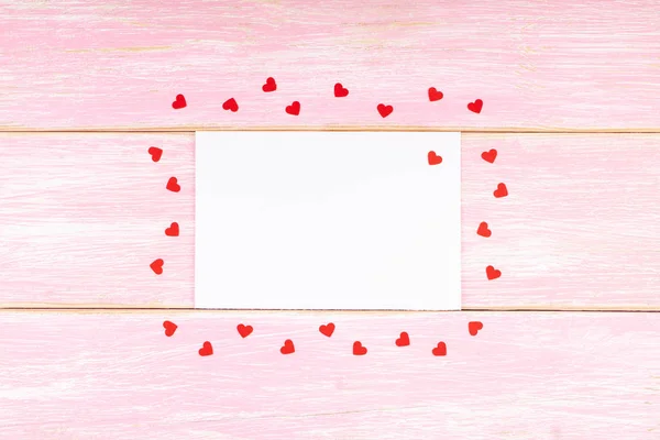 Tarjeta blanca en blanco con corazones rojos de confeti sobre fondo de madera rosa — Foto de Stock
