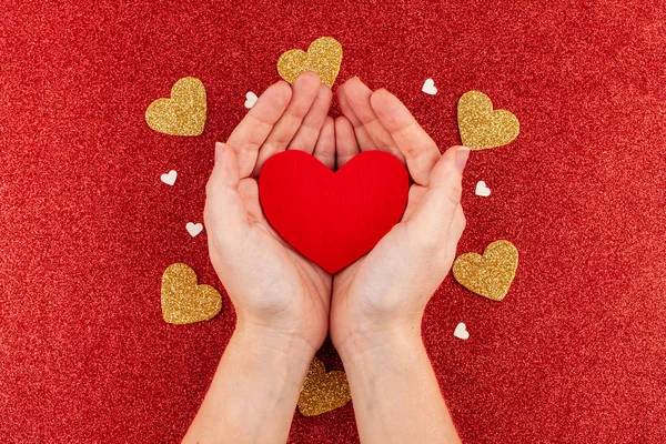 Manos de mujer sosteniendo el corazón rojo sobre fondo rojo brillo con corazones de oro — Foto de Stock