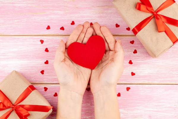 Žena ruce drží červené srdce na růžové dřevěné pozadí s červenými konfetti srdce — Stock fotografie
