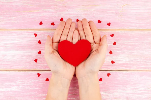 Woman Hands Holding Red Heart on Pink Wooden Background with Red Confetti Hearts — Stock Photo, Image