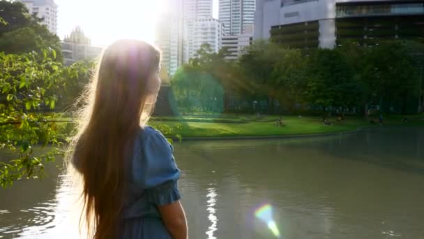 Mooi gelukkig meisje staande door meer in park met bomen in de zonsondergang zonnestralen — Stockvideo