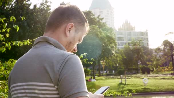 Hombre serio usando su teléfono inteligente en el parque público con rascacielos en segundo plano — Vídeo de stock